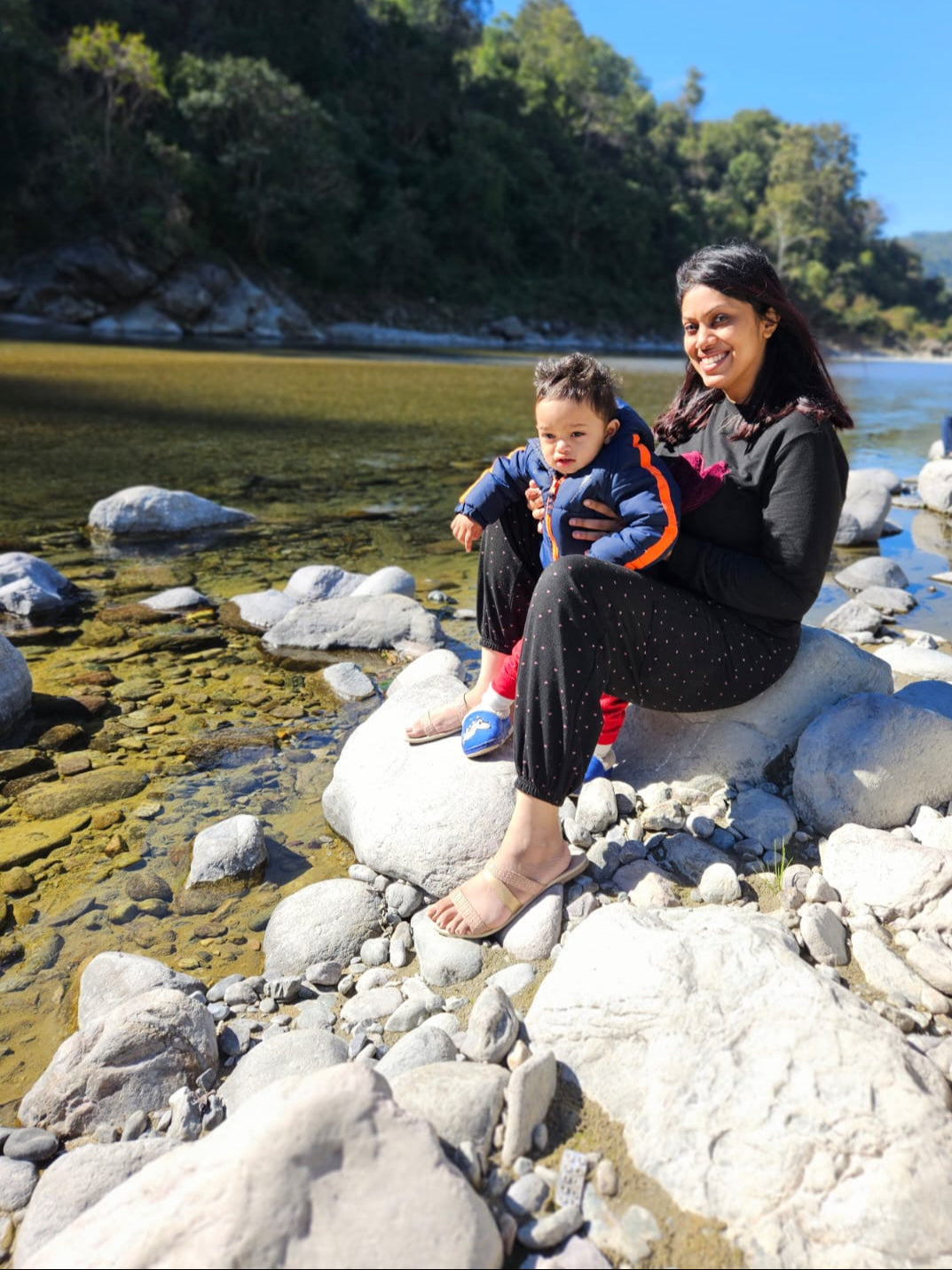 Woman holding a child on rocks by a river with trees in the background